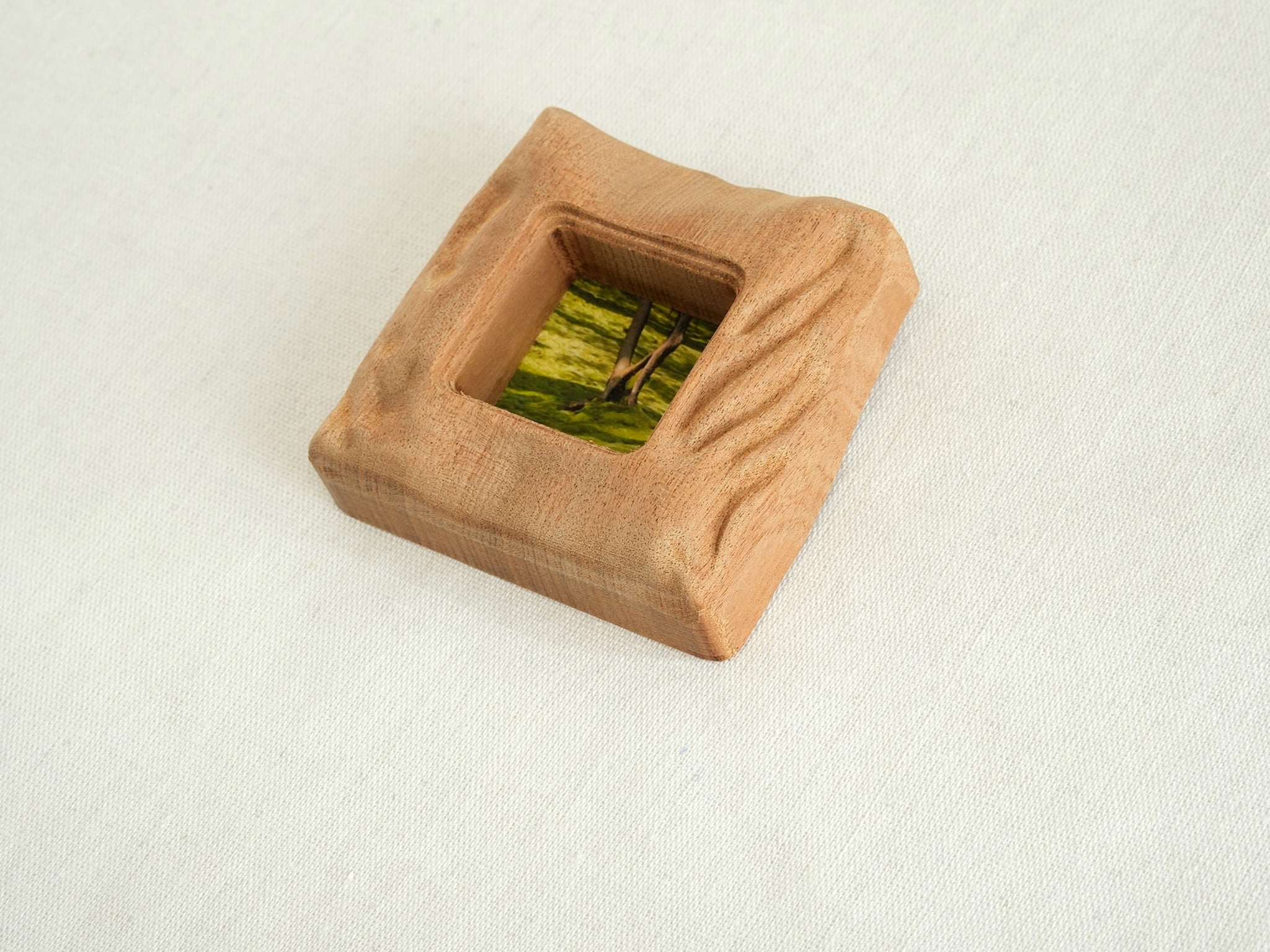 A left-side isometric view of the handmade mini solid wood frame, emphasizing the organic, uneven hand-carved surface and the smooth, rounded corners. The natural grain of the Sapele wood flows seamlessly across the tactile, sculpted edges.