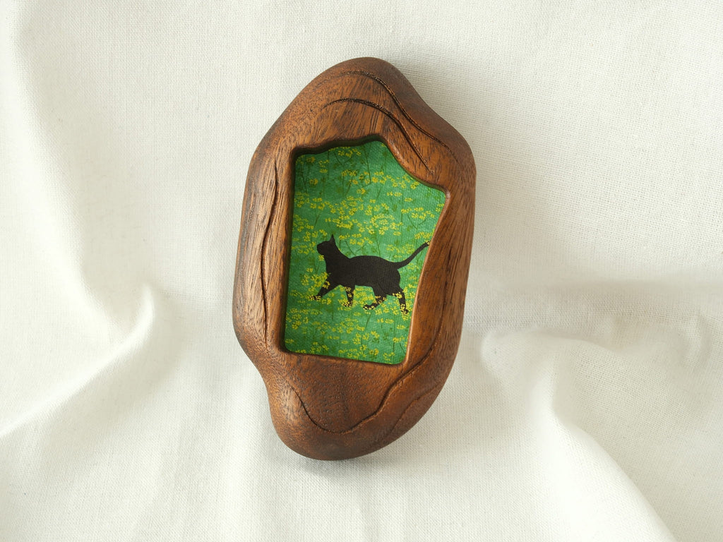 A lifestyle shot of the irregular organic wood frame leaning against a soft white fabric, displaying its thick sculptural profile and the vibrant artwork of a black cat in a yellow flower field.