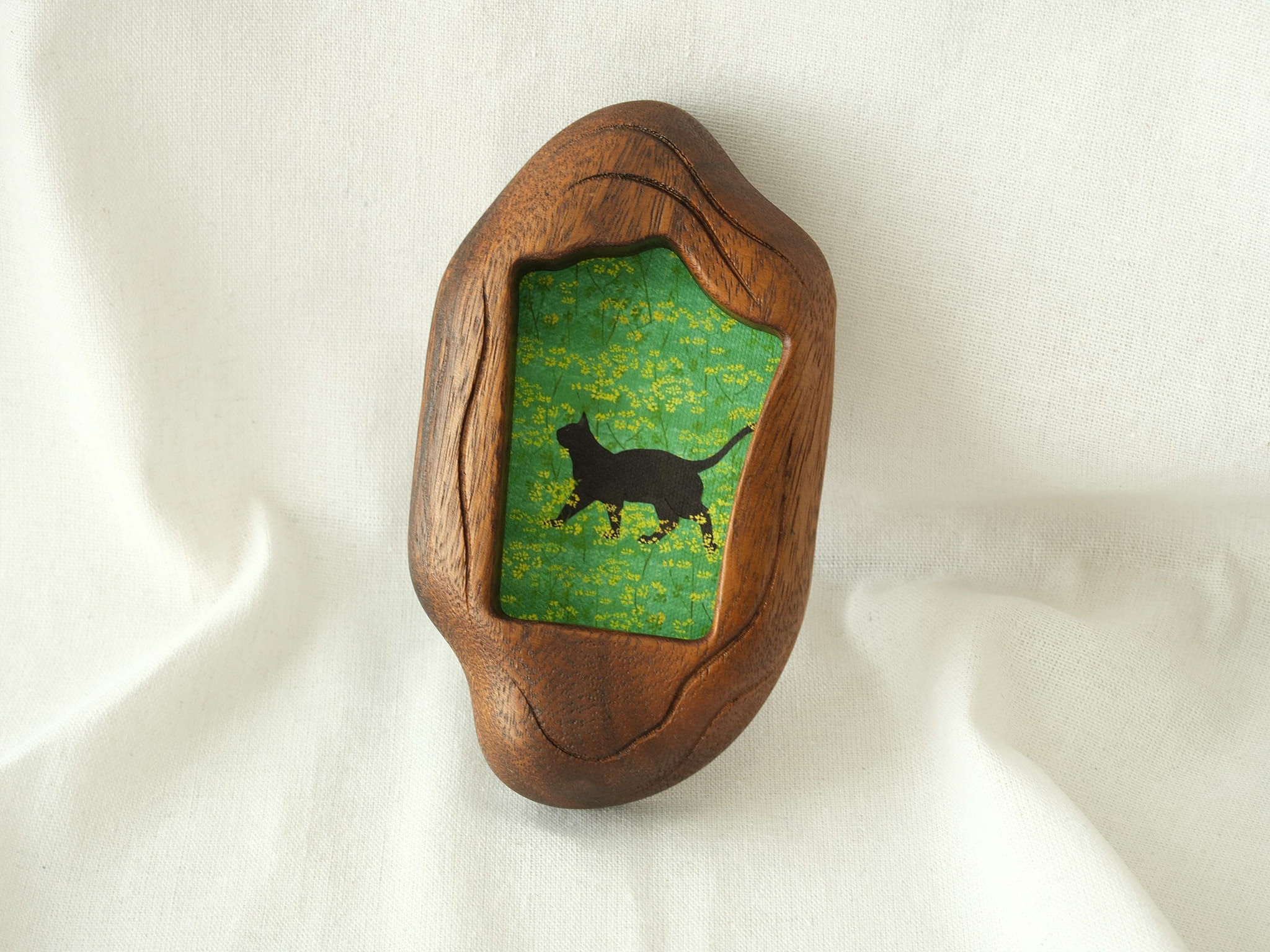 A lifestyle shot of the irregular organic wood frame leaning against a soft white fabric, displaying its thick sculptural profile and the vibrant artwork of a black cat in a yellow flower field.