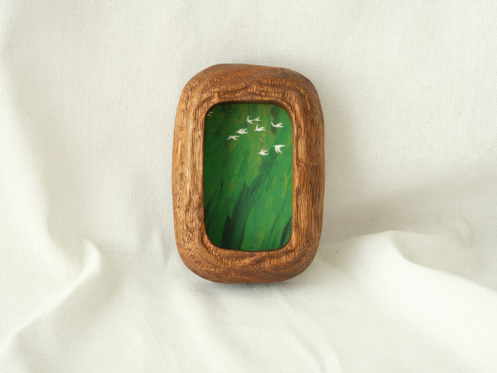 A vertical lifestyle shot of the Rounded Rectangle solid wood frame leaning against a soft white background, showcasing its thick timber profile and the vibrant green tones of the forest birds artwork.