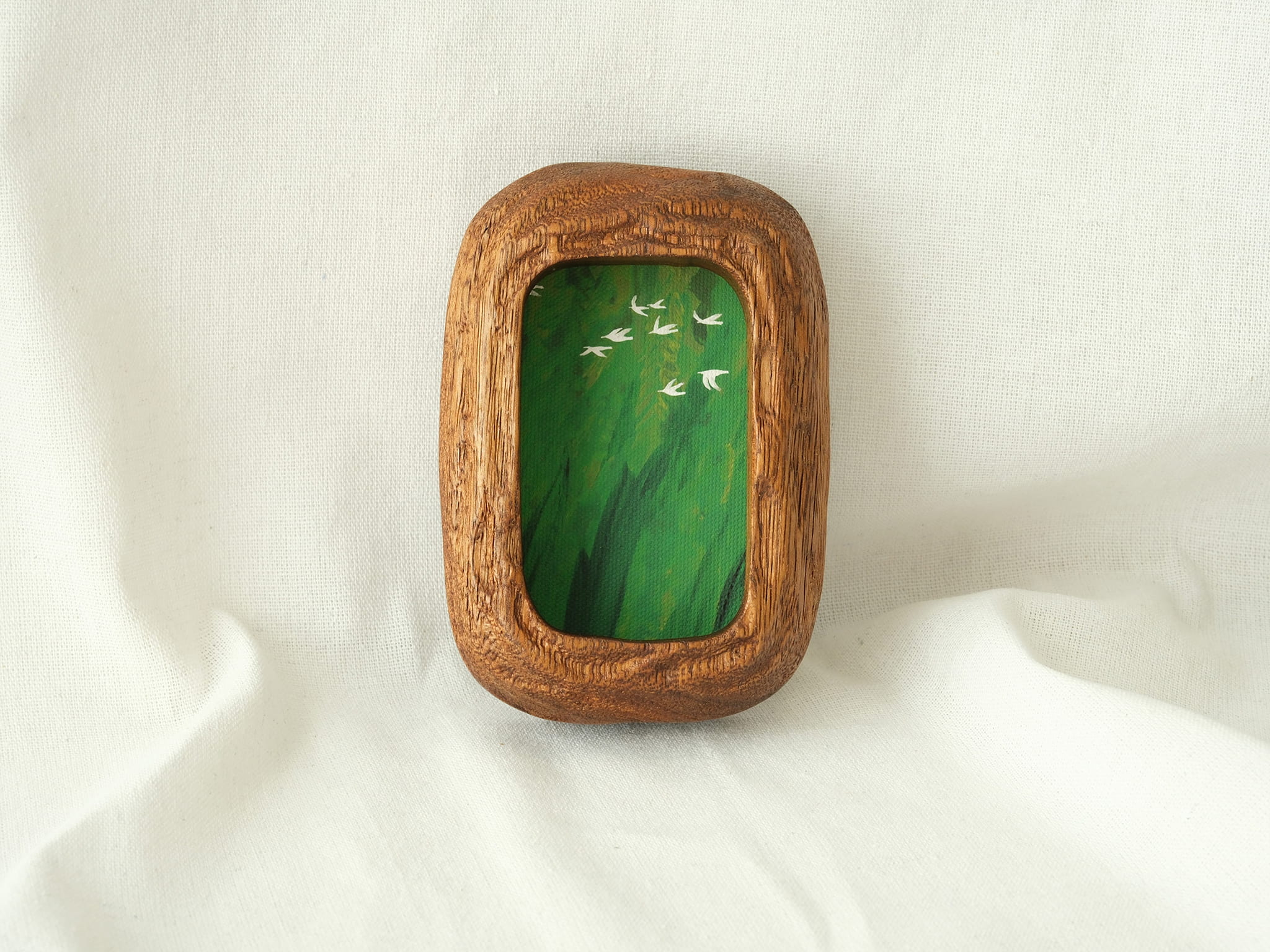 A vertical lifestyle shot of the Rounded Rectangle solid wood frame leaning against a soft white background, showcasing its thick timber profile and the vibrant green tones of the forest birds artwork.