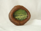 A perspective three-quarter view of the lake-shaped organic solid wood frame standing upright, showcasing the thick handcrafted wood contours and the serene landscape painting of a green meadow filled with wildflowers.