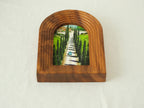 A high-angle three-quarter view from the front, showcasing the arched "Tuscany" solid wood frame standing upright, revealing the deep stepped wood contours and the serene oil-style painting of the cypress avenue.