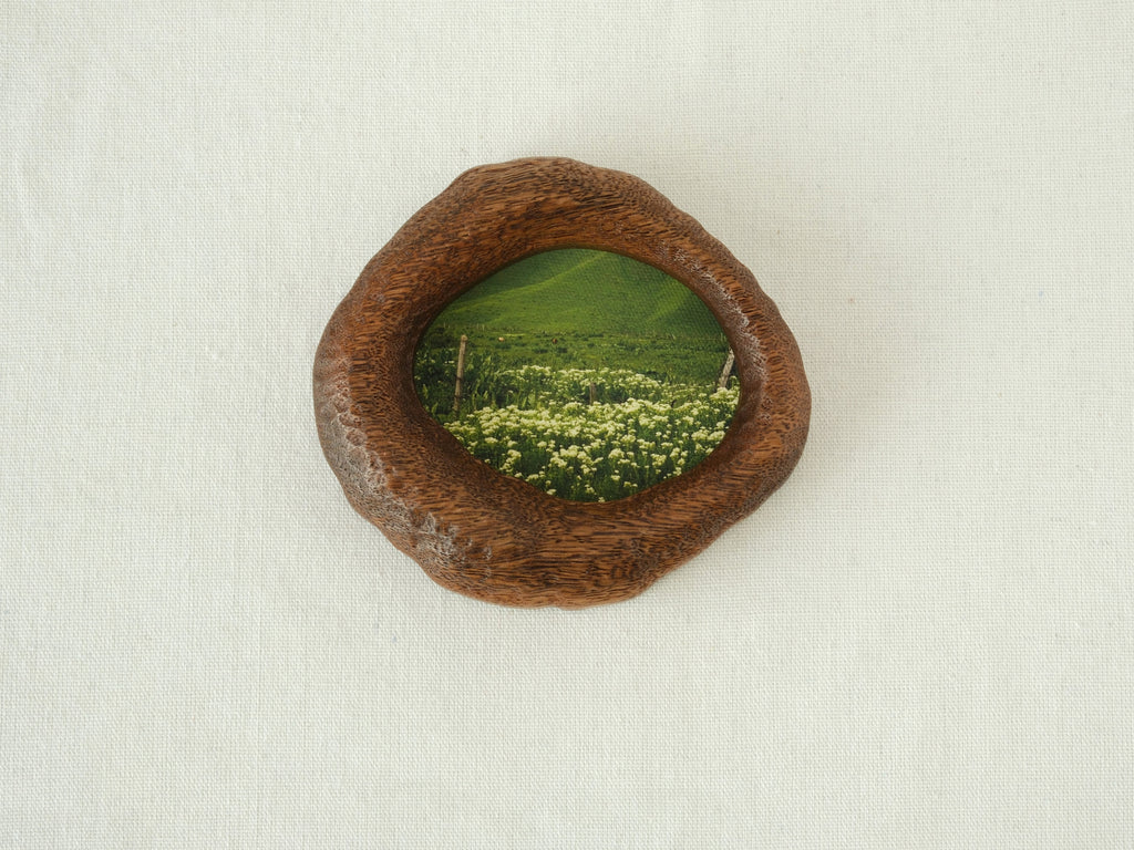 Front view of the lake-shaped organic solid wood frame, featuring a serene landscape painting of a green meadow filled with white wildflowers.