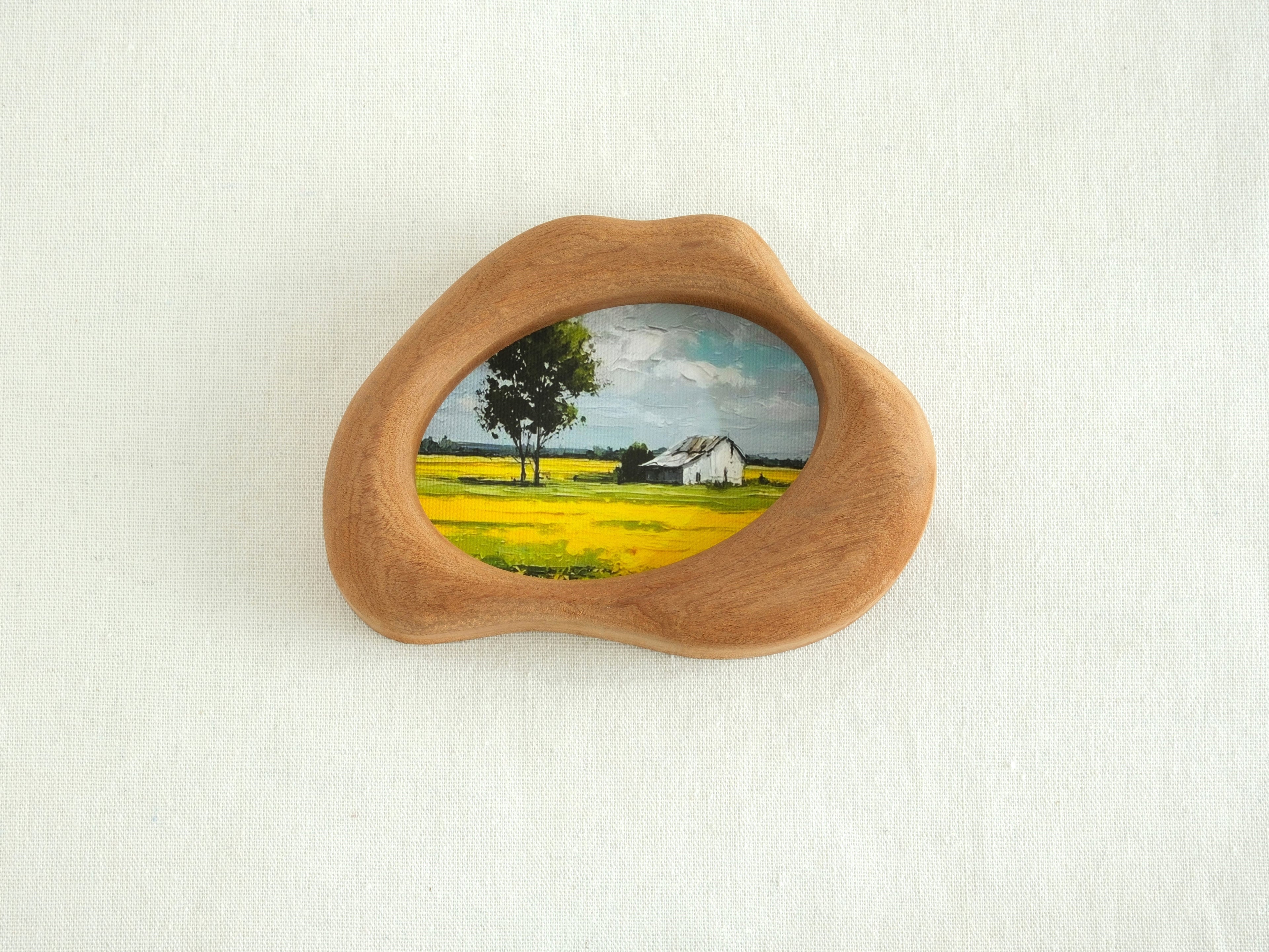 A front view of the handmade solid wood art frame featuring a soft, organic cloud-shaped silhouette. The smooth, hand-sculpted curves of the wood encase a landscape painting, creating the aesthetic of a floating piece of nature.