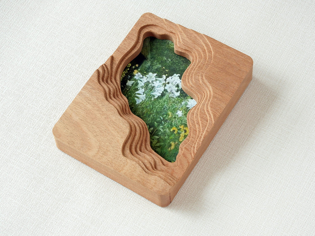 Solid wood wall art picture frame with flowing stream-shaped cutout and floral meadow artwork, high-angle side view.
