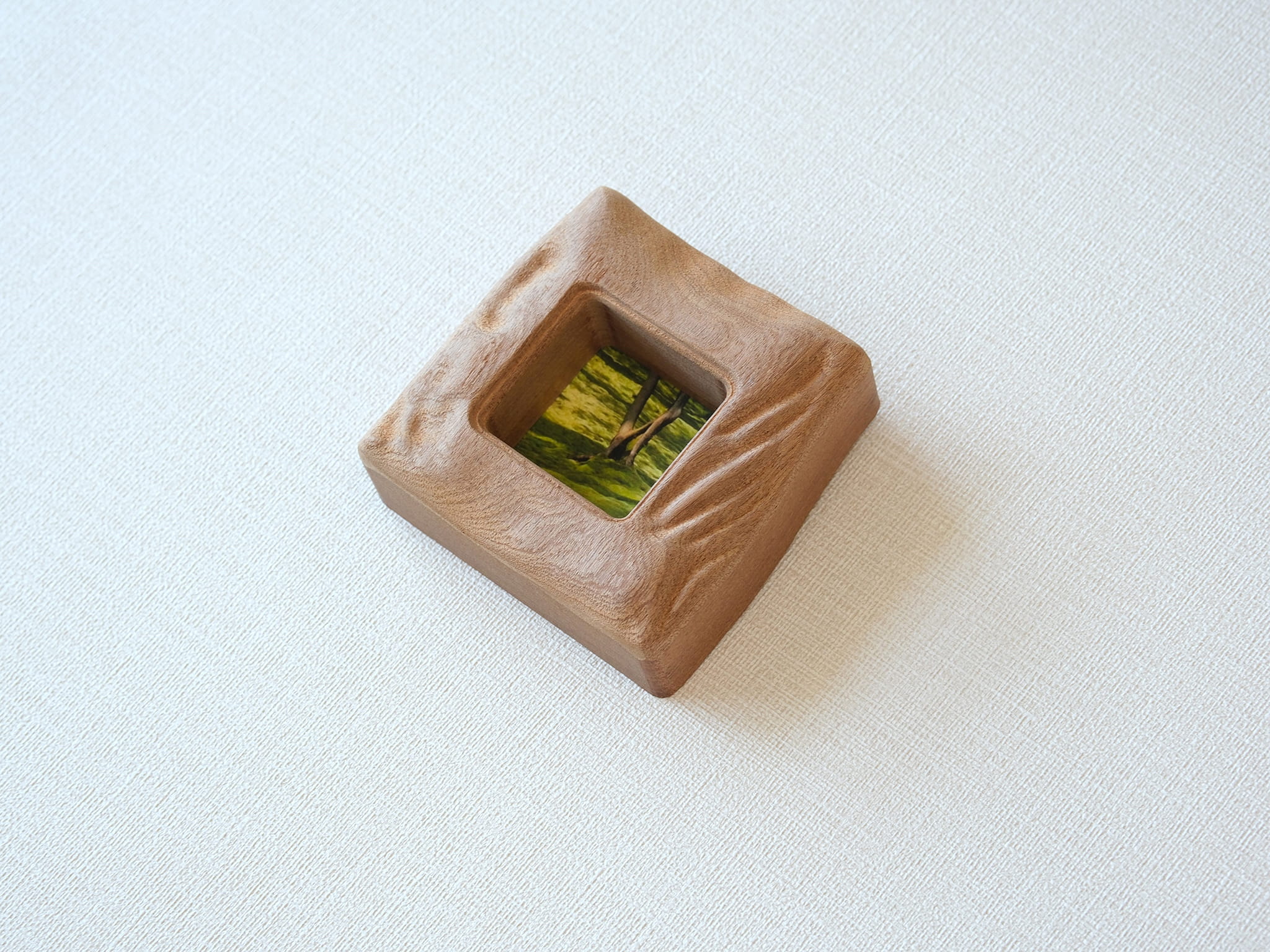 A left-side isometric view of the handmade mini solid wood frame, emphasizing the organic, uneven hand-carved surface and the smooth, rounded corners. The natural grain of the Sapele wood flows seamlessly across the tactile, sculpted edges.