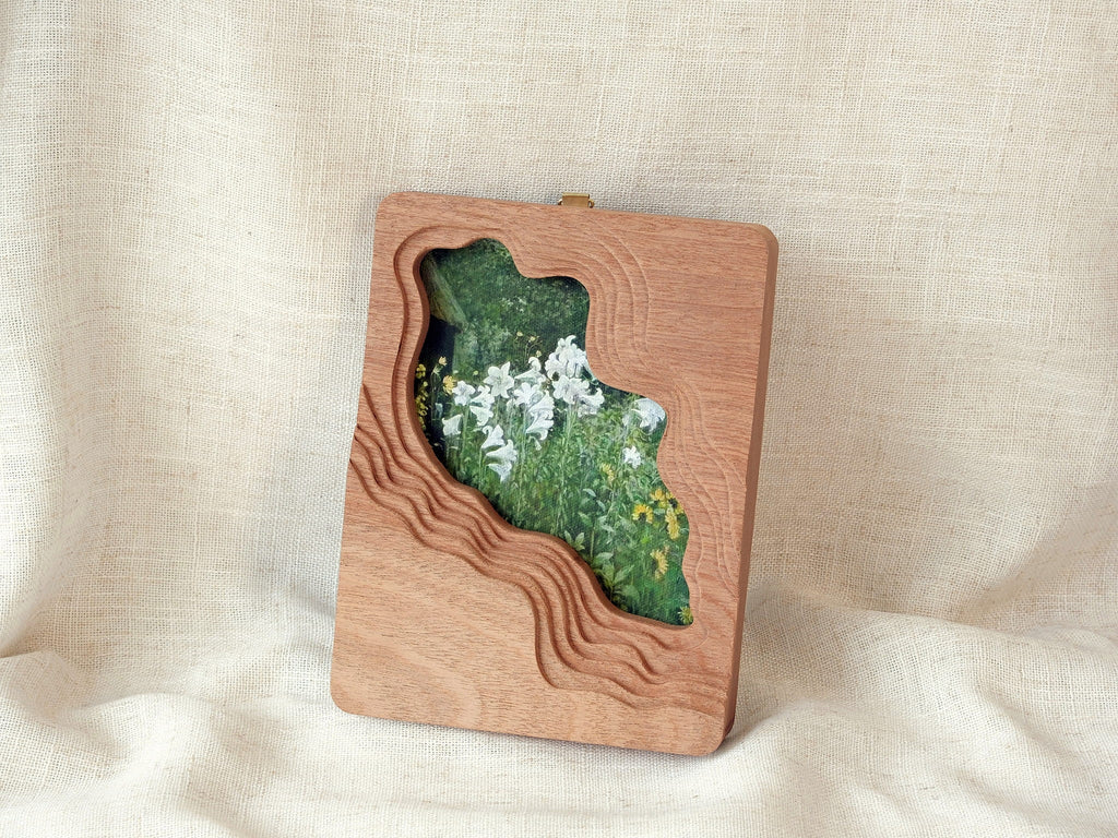 Large solid wood wall hanging art frame with flowing stream-shaped cutout and floral meadow artwork, leaning display.