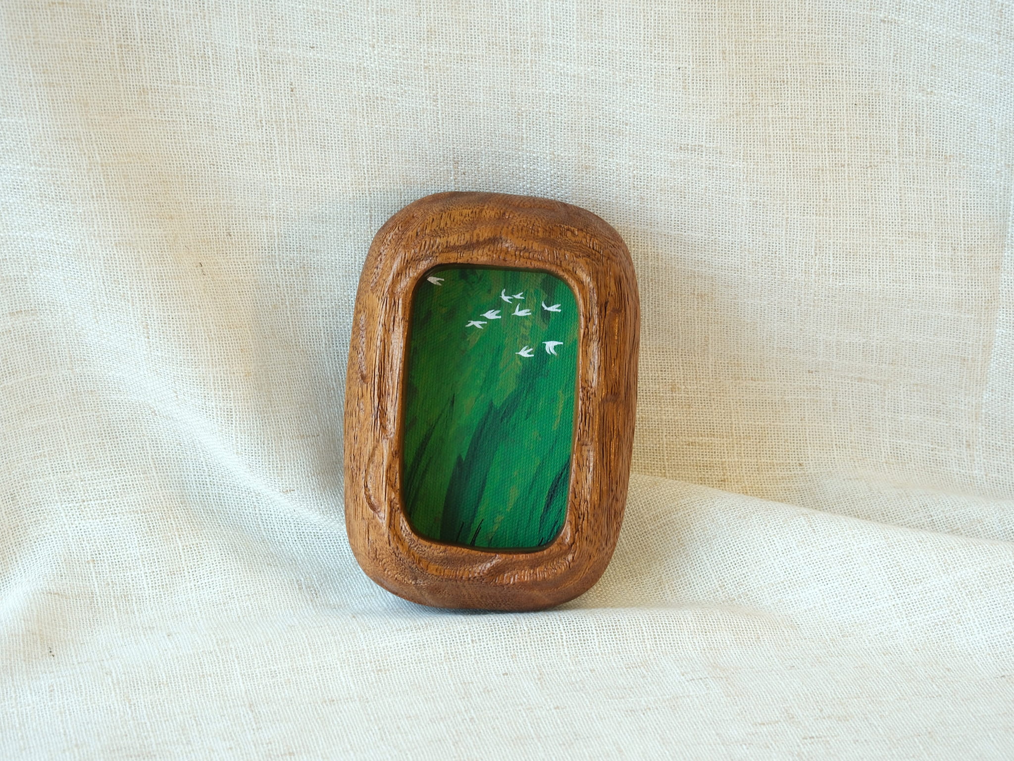 A vertical lifestyle shot of the Rounded Rectangle solid wood frame leaning against a soft white background, showcasing its thick timber profile and the vibrant green tones of the forest birds artwork.