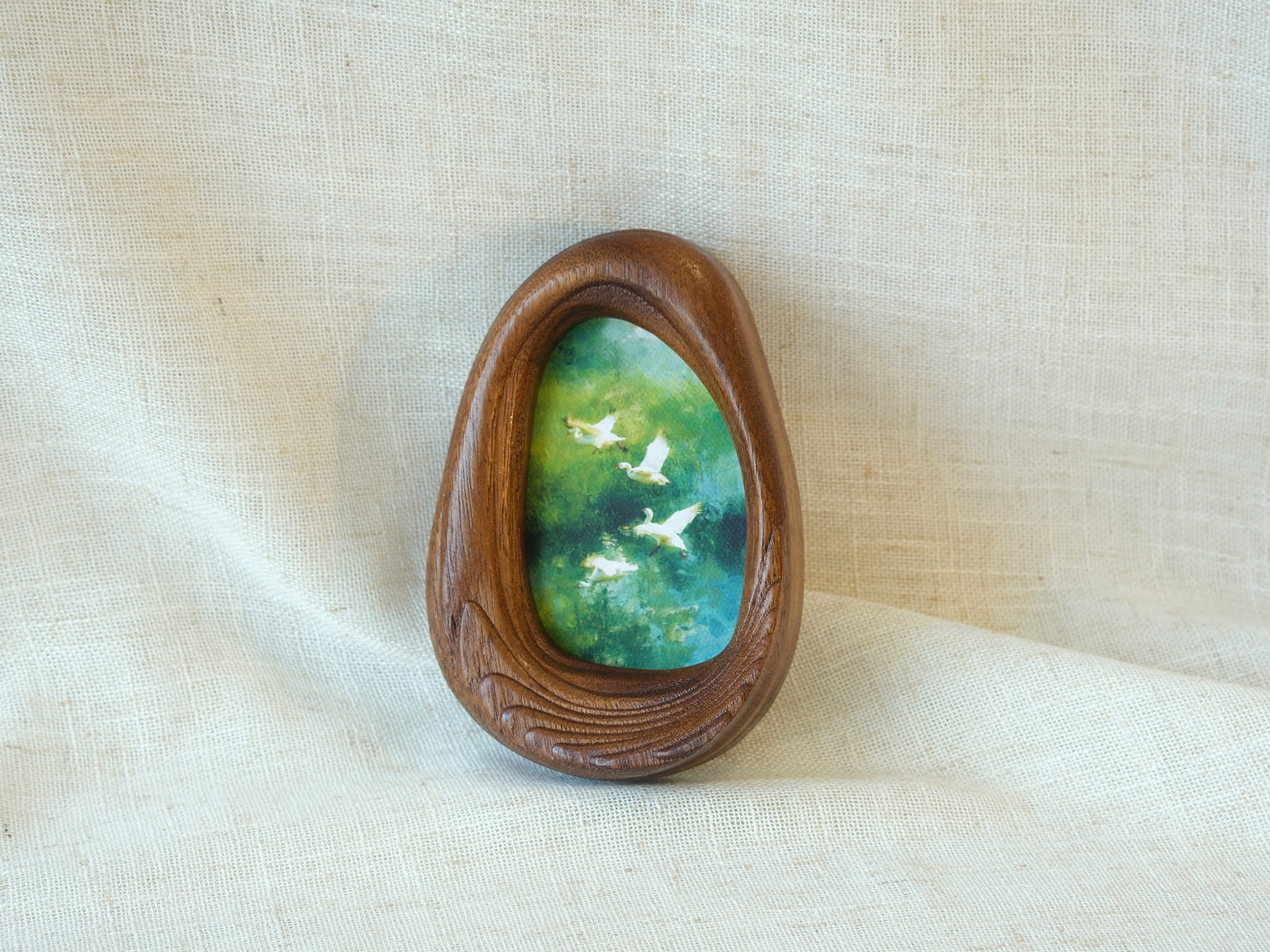 A realistic lifestyle shot of the Waterdrop solid wood frame leaning against a soft white background, displaying the vibrant colors of the flying birds and the rich, dark tones of the natural wood.