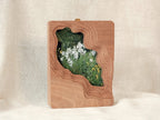 Handmade solid wood wall art picture frame with flowing stream-shaped cutout and floral meadow artwork, standing display.