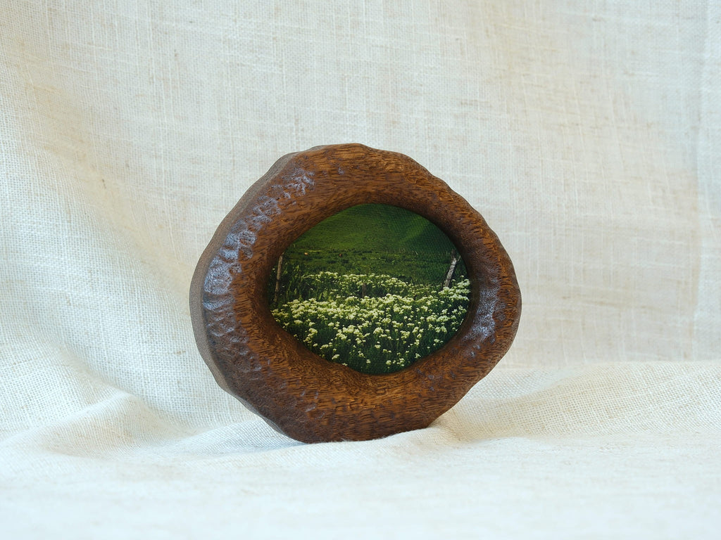 A perspective three-quarter view of the lake-shaped organic solid wood frame standing upright, showcasing the thick handcrafted wood contours and the serene landscape painting of a green meadow filled with wildflowers.
