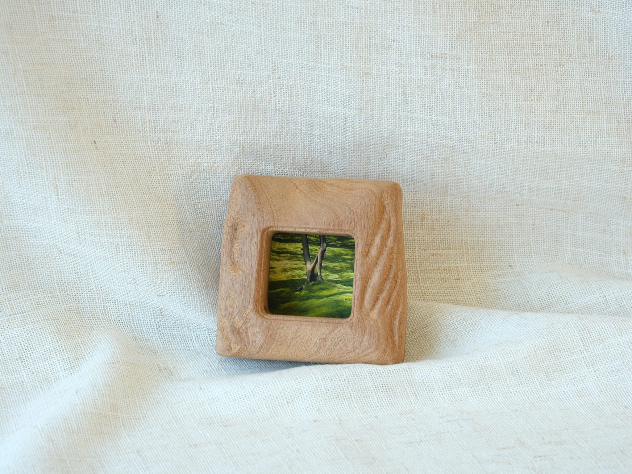 A direct front view of the mini solid wood frame leaning gently against a plain white wall. The natural daylight accentuates the deeply sculpted, fluid "Moderno Organico" wavy patterns on the frame's right side, casting subtle shadows.