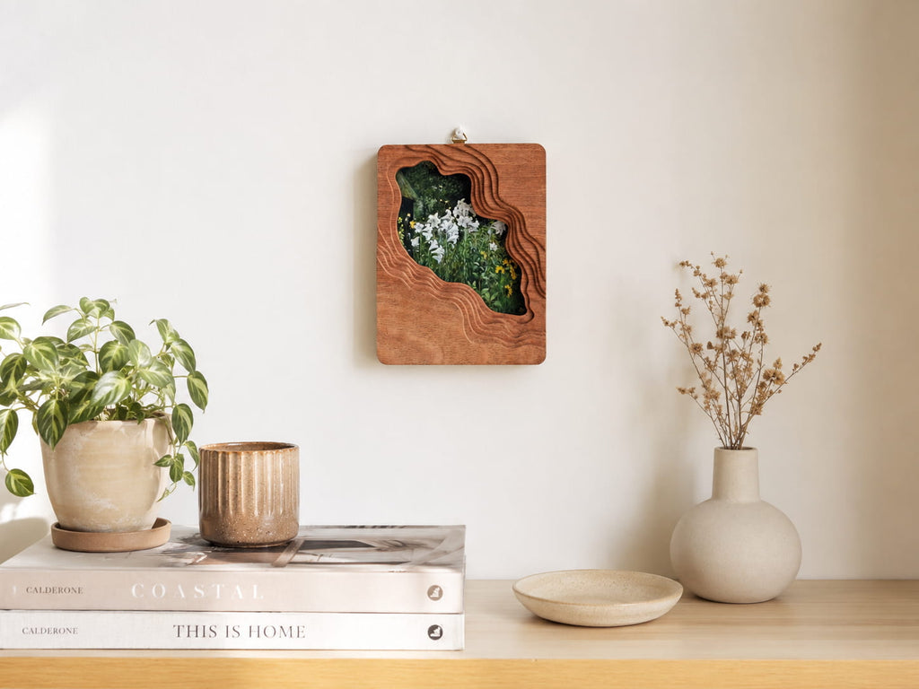 Large solid wood wall hanging art frame with floral meadow artwork displayed on a white wall above a wooden table.