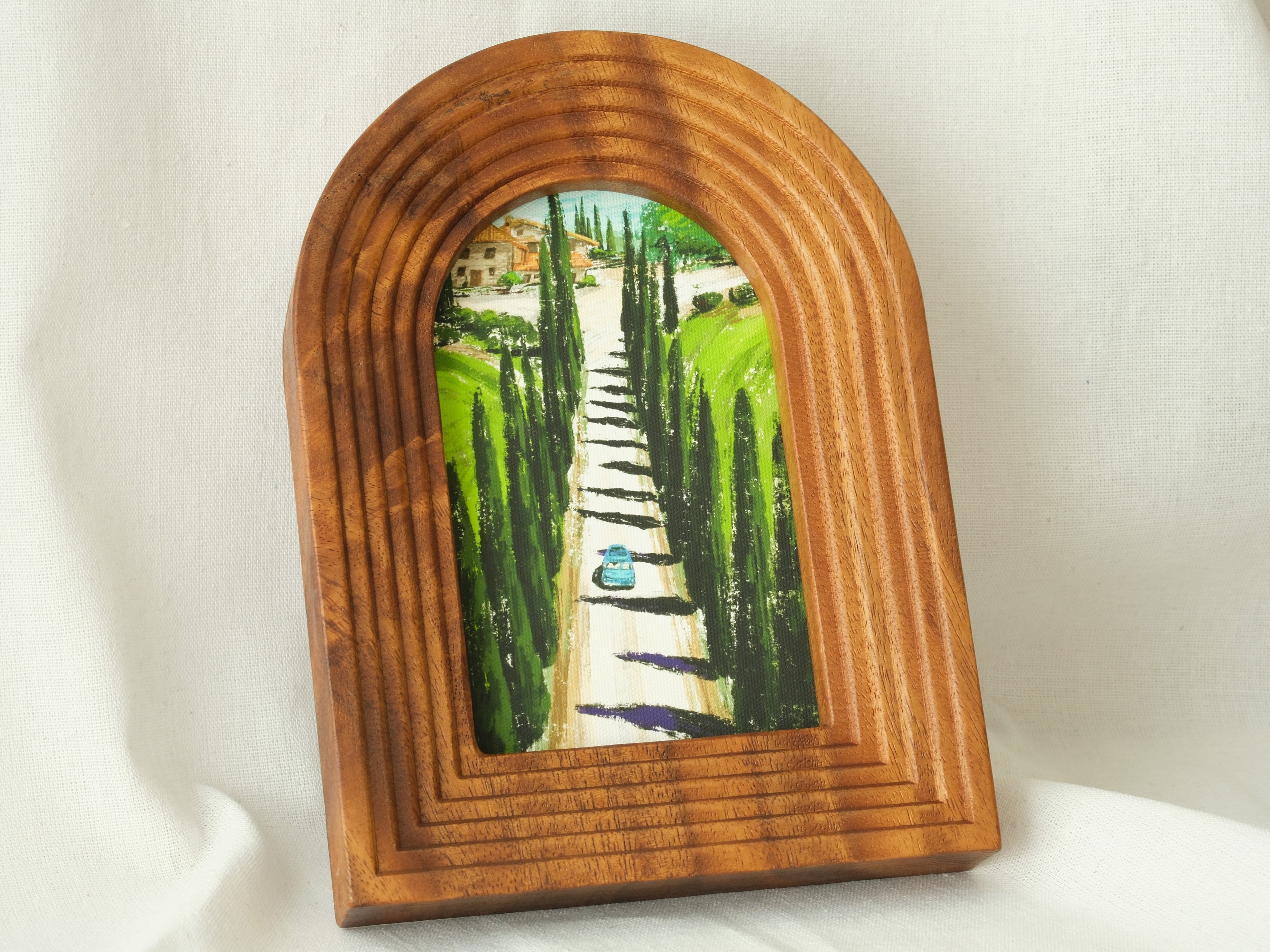 A soft-focus lifestyle shot of the arched "Tuscany" solid wood frame resting against a gently draped white fabric, showcasing its sculptural form and the serene oil-style painting of the cypress avenue.