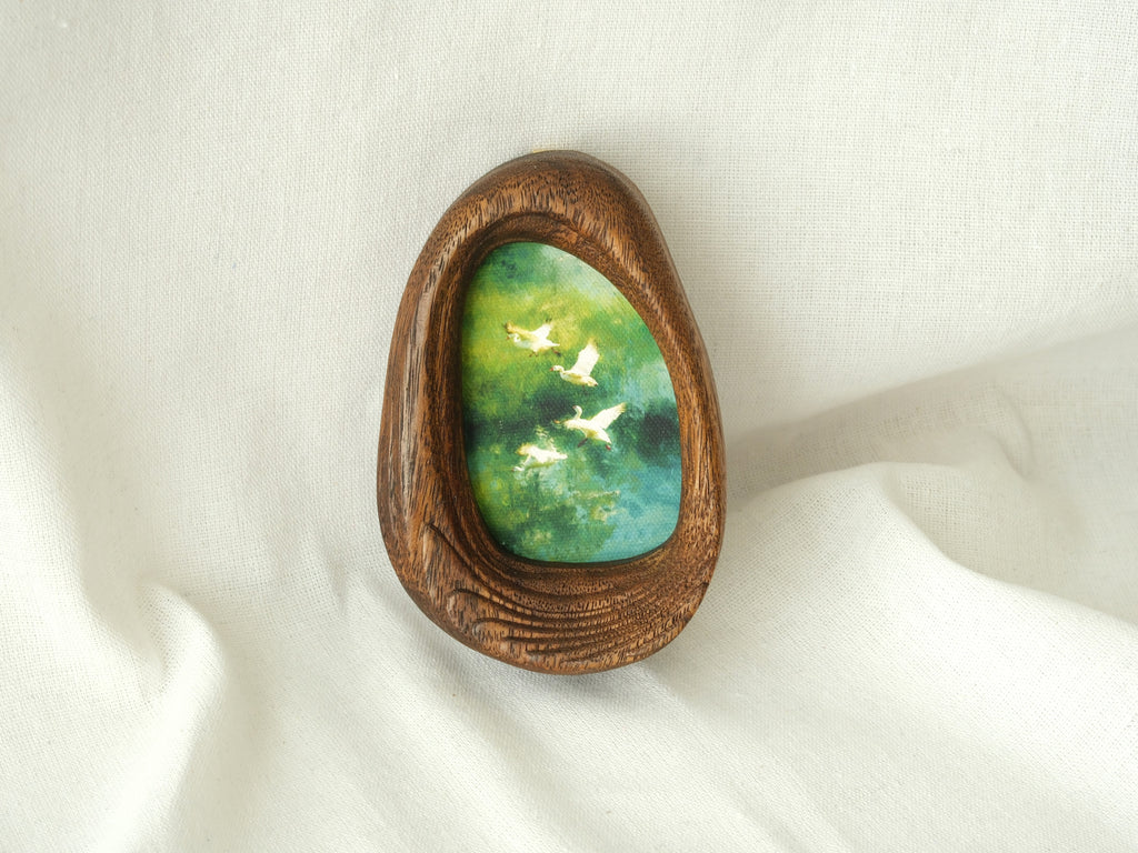 A realistic lifestyle shot of the Waterdrop solid wood frame leaning against a soft white background, displaying the vibrant colors of the flying birds and the rich, dark tones of the natural wood.