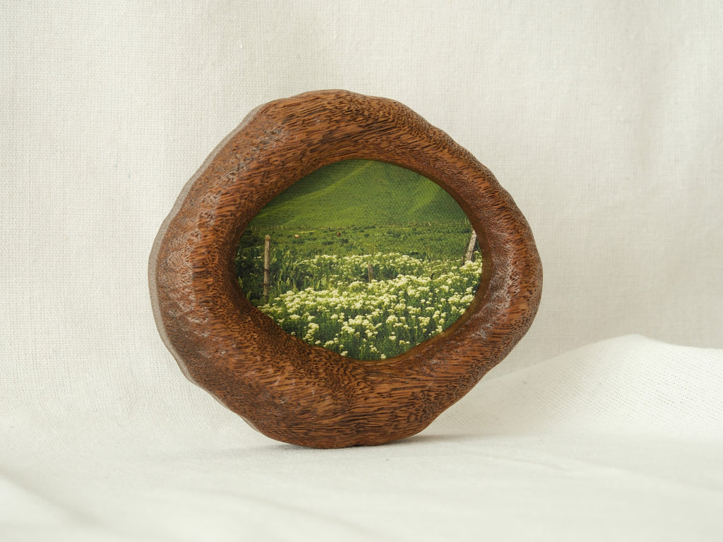 A perspective three-quarter view of the lake-shaped organic solid wood frame standing upright, showcasing the thick handcrafted wood contours and the serene landscape painting of a green meadow filled with wildflowers.