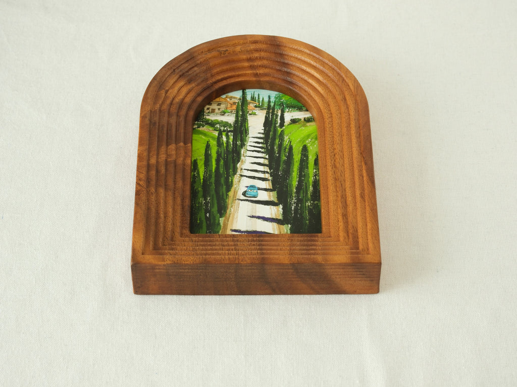 A high-angle three-quarter view from the front, showcasing the arched "Tuscany" solid wood frame standing upright, revealing the deep stepped wood contours and the serene oil-style painting of the cypress avenue.