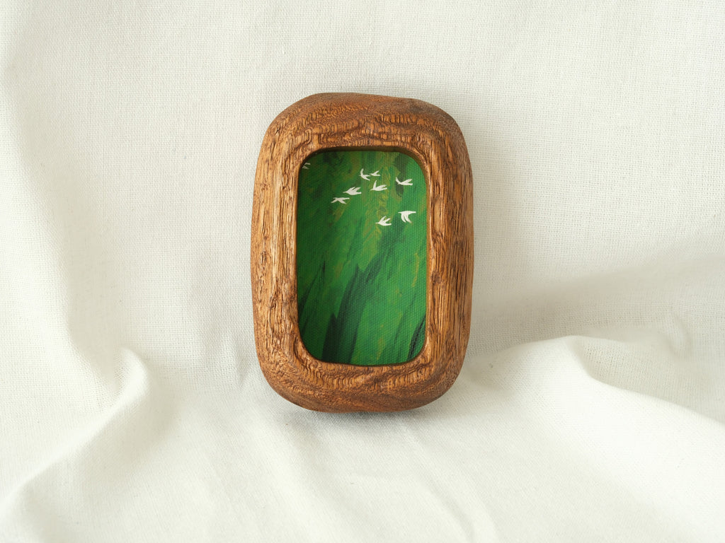 A vertical lifestyle shot of the Rounded Rectangle solid wood frame leaning against a soft white background, showcasing its thick timber profile and the vibrant green tones of the forest birds artwork.