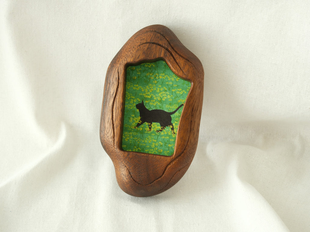 A lifestyle shot of the irregular organic wood frame leaning against a soft white fabric, displaying its thick sculptural profile and the vibrant artwork of a black cat in a yellow flower field.