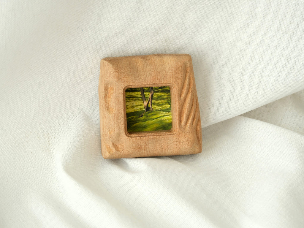A direct front view of the mini solid wood frame leaning gently against a plain white wall. The natural daylight accentuates the deeply sculpted, fluid "Moderno Organico" wavy patterns on the frame's right side, casting subtle shadows.