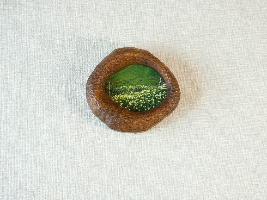 Front view of the lake-shaped organic solid wood frame, featuring a serene landscape painting of a green meadow filled with white wildflowers.