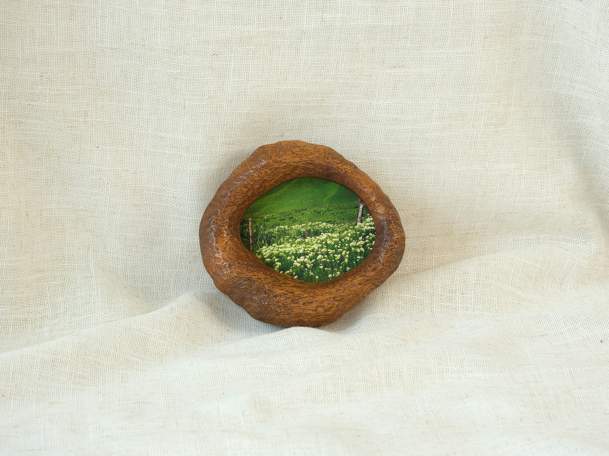 A soft-focus lifestyle shot of the lake-shaped organic wood frame resting on drapes of white fabric, showcasing its sculptural presence as a gentle wall accent with the meadow landscape art.