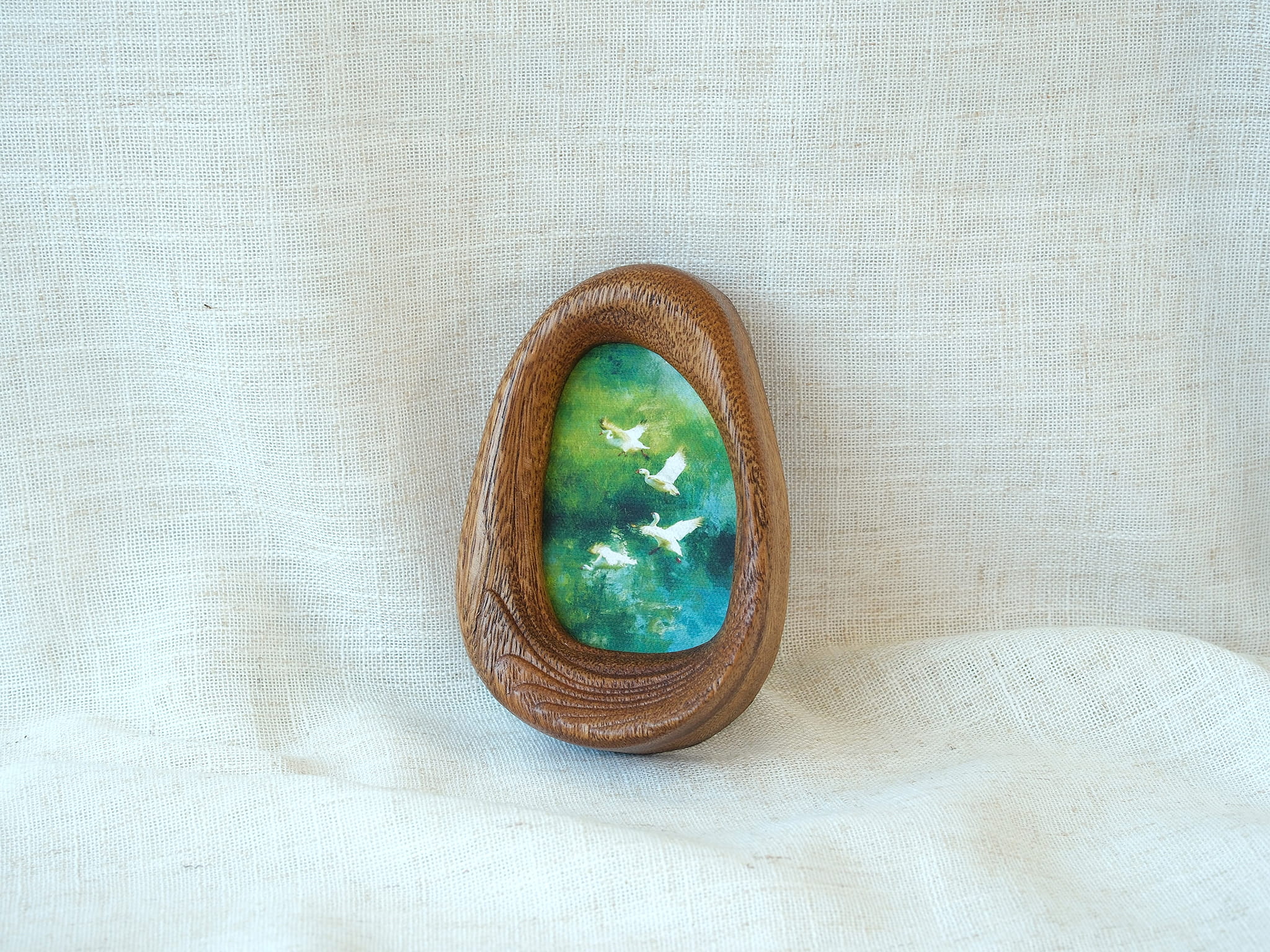 A realistic lifestyle shot of the Waterdrop solid wood frame leaning against a soft white background, displaying the vibrant colors of the flying birds and the rich, dark tones of the natural wood.