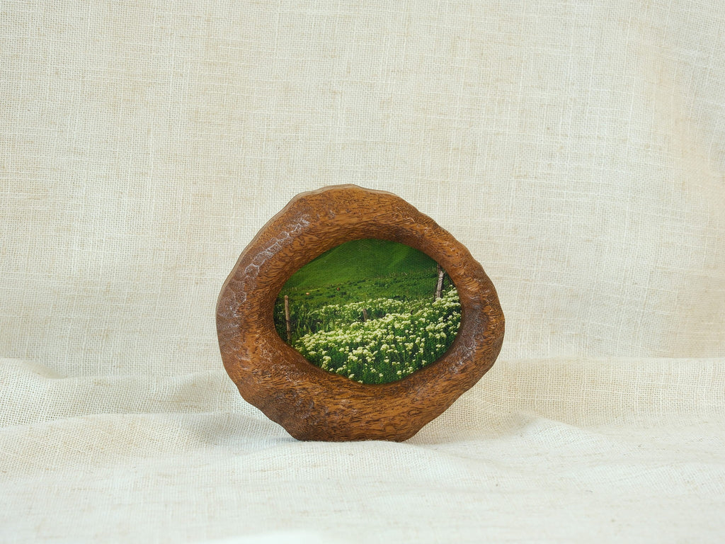A perspective three-quarter view of the lake-shaped organic solid wood frame standing upright, showcasing the thick handcrafted wood contours and the serene landscape painting of a green meadow filled with wildflowers.