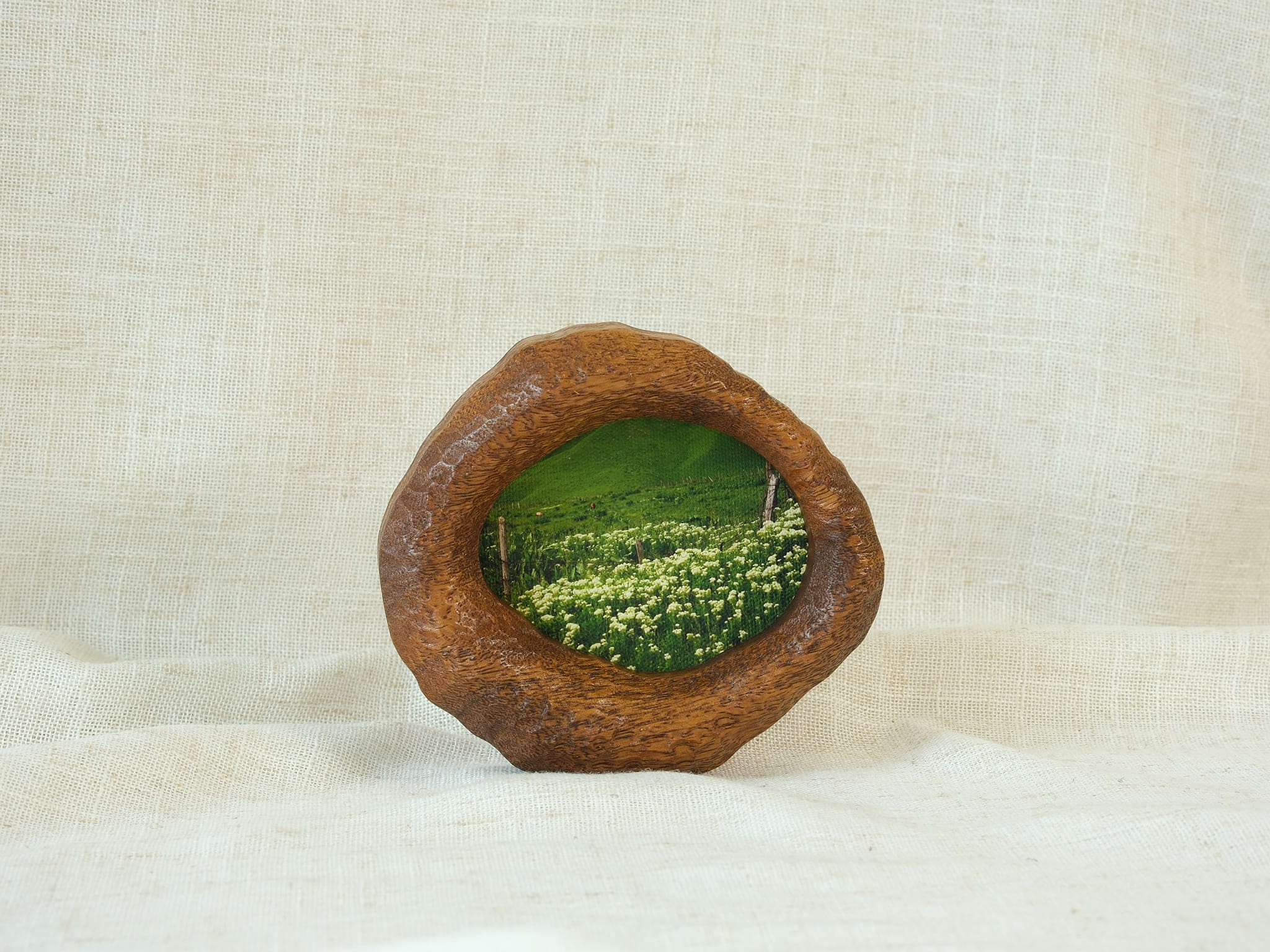 A perspective three-quarter view of the lake-shaped organic solid wood frame standing upright, showcasing the thick handcrafted wood contours and the serene landscape painting of a green meadow filled with wildflowers.