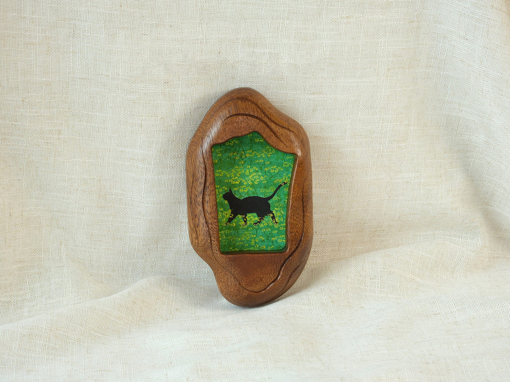 A lifestyle shot of the irregular organic wood frame leaning against a soft white fabric, displaying its thick sculptural profile and the vibrant artwork of a black cat in a yellow flower field.