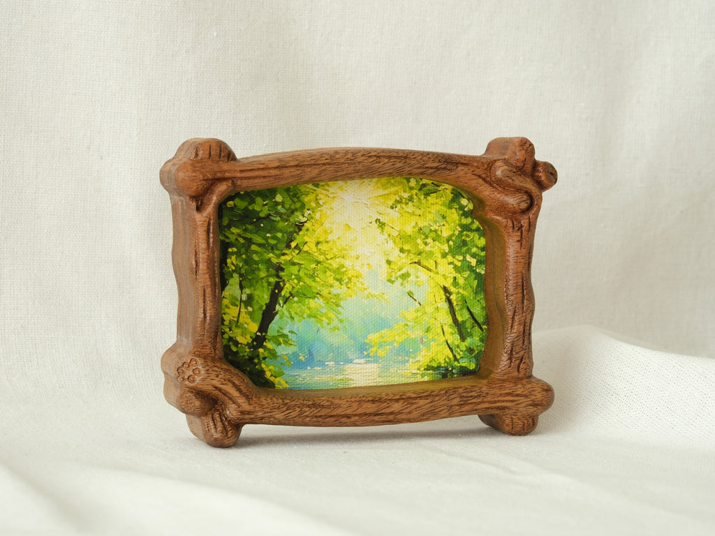 A side-angle view of a handcrafted wood frame with an organic branch-like texture, featuring a vibrant forest sunlight oil painting.